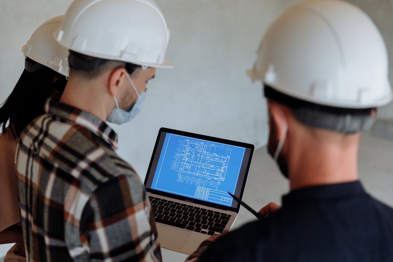 Engineers wearing hard hats and masks review a construction blueprint on a laptop indoors.